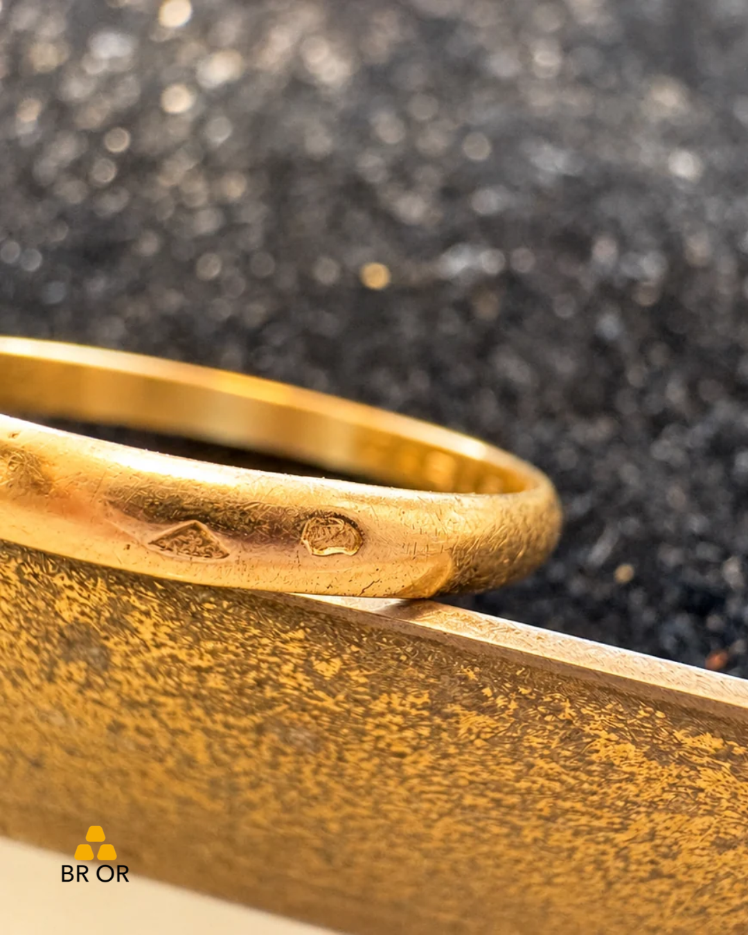 Détail de deux poinçons sur une bague dorée Deux poinçons, l’un losangique et l’autre arrondi, sur une bague dorée posée sur un support métallique. Logo BR OR en bas à gauche.